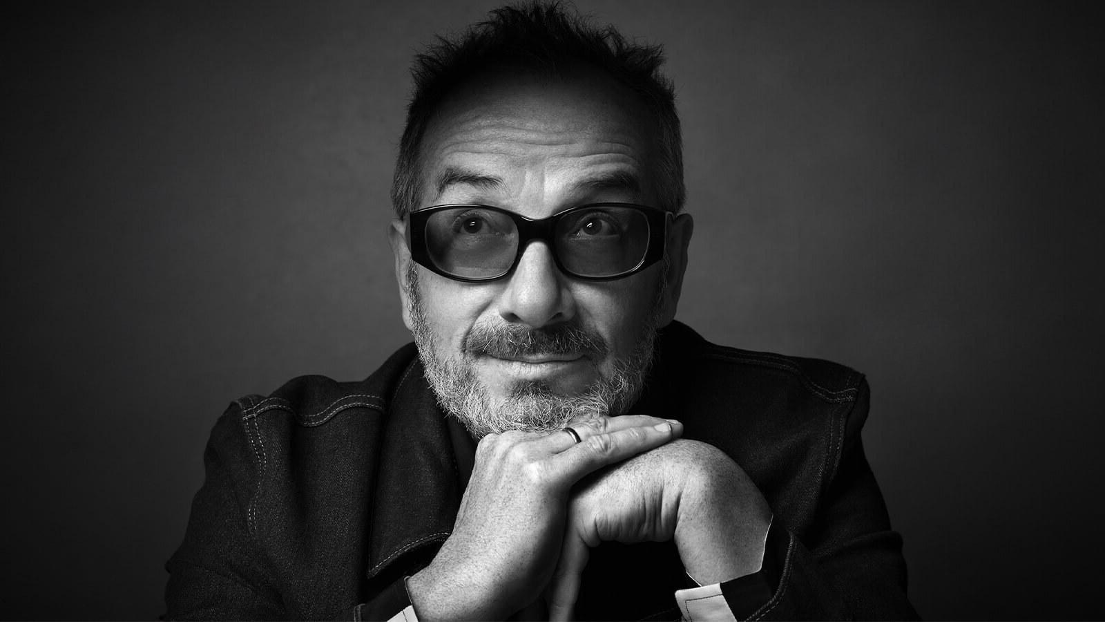 A black and white portrait of singer, songwriter, and producer Elvis Costello wearing dark-tinted glasses and a wedding band.