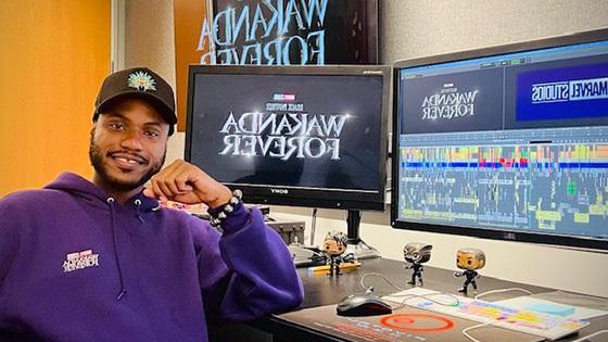 Alain sits at a desk with multiple monitors that read “Wakanda Forever” and “Marvel Studios.” He is wearing a purple hoodie.