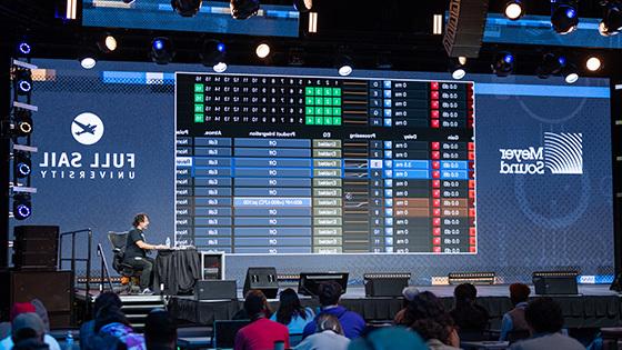 A person is seated at a desk on a stage in front of a crowd of students while a large LED screen behind displays a mirror of the audio program running on their computer.