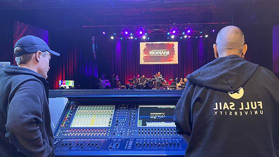 Two people's backs are facing the camera as they overlook an audio console and stage at The Plaza Live.