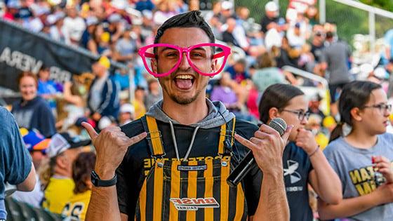 A man standing and looking excited while holding a microphone in oversized pink glasses and yellow and black striped overalls in the foreground of a large crowd at a stadium.