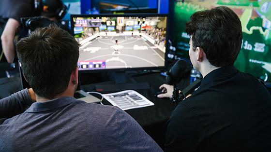 Two people have their backs to the camera while watching ‘NBA 2K’ gameplay on a small monitor on a table in front of them.