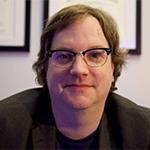 Up close headshot of a man wearing glasses and a black shirt against a wall with framed degrees.