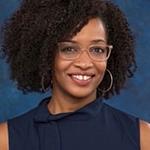 Tiana, an African American woman, sits smiling at the camera. She is wearing a sleeveless navy blue blouse, silver hoop earrings, and glasses with translucent beige frames.